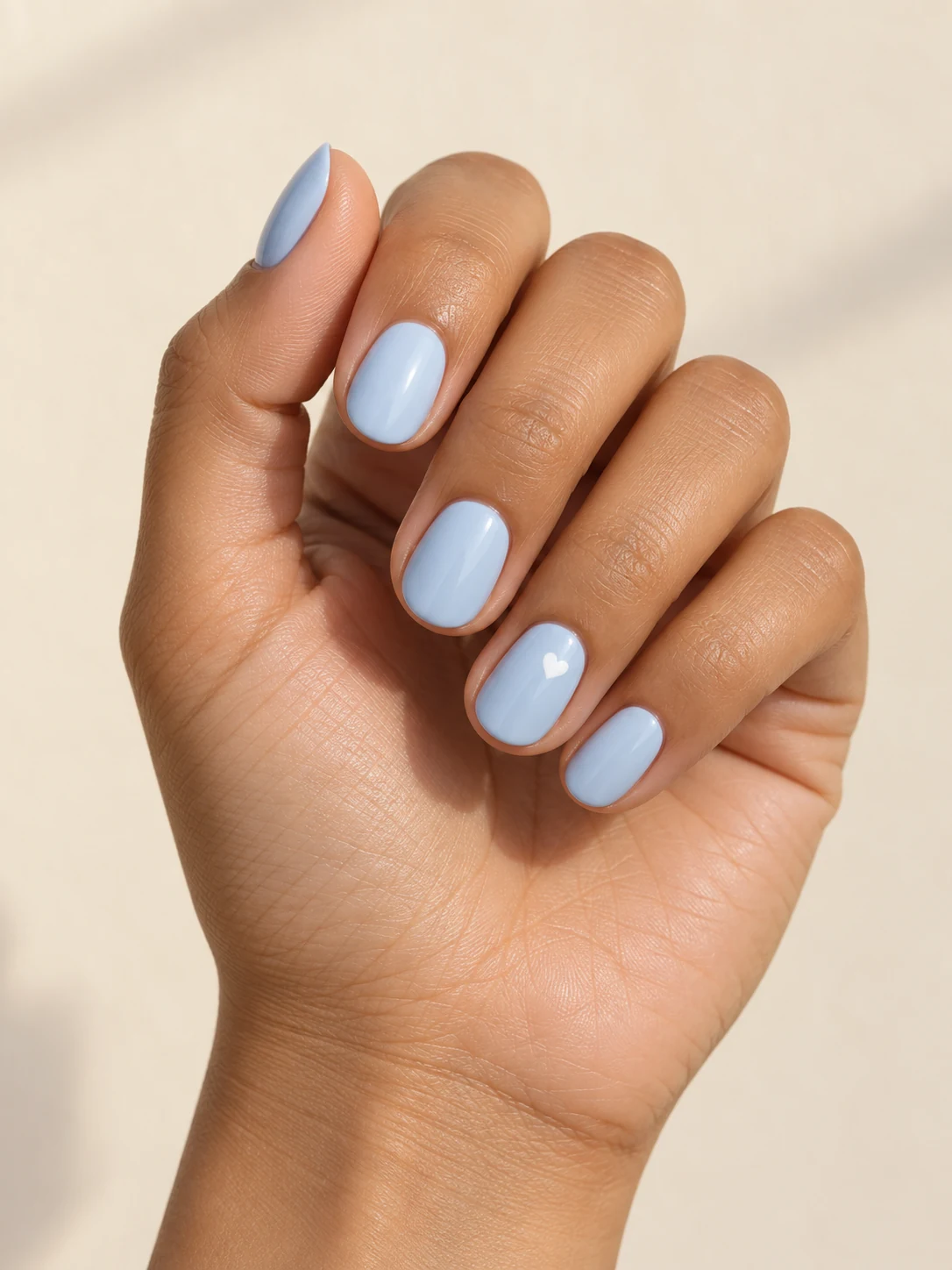 Pale powder-blue nails with a single tiny white heart on one ring-finger accent, restrained glossy finish on warm medium skin; short oval manicure.
