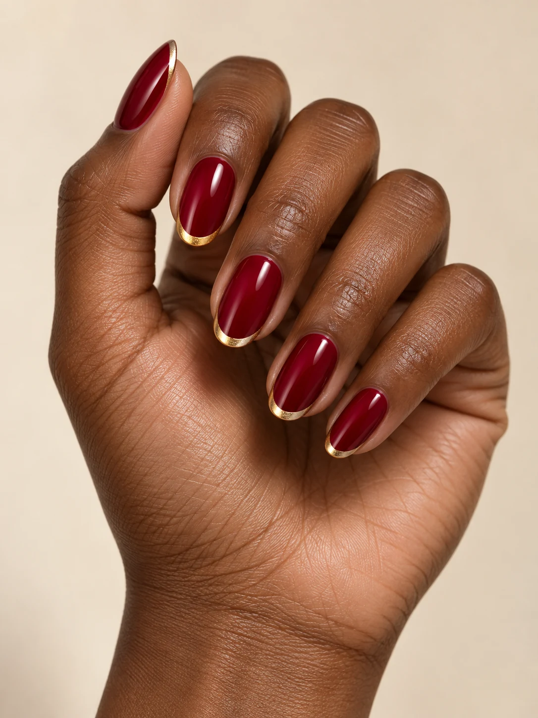 Deep cranberry-red nails with narrow metallic gold French tips, restrained festive gloss on deep warm brown skin; medium oval manicure.