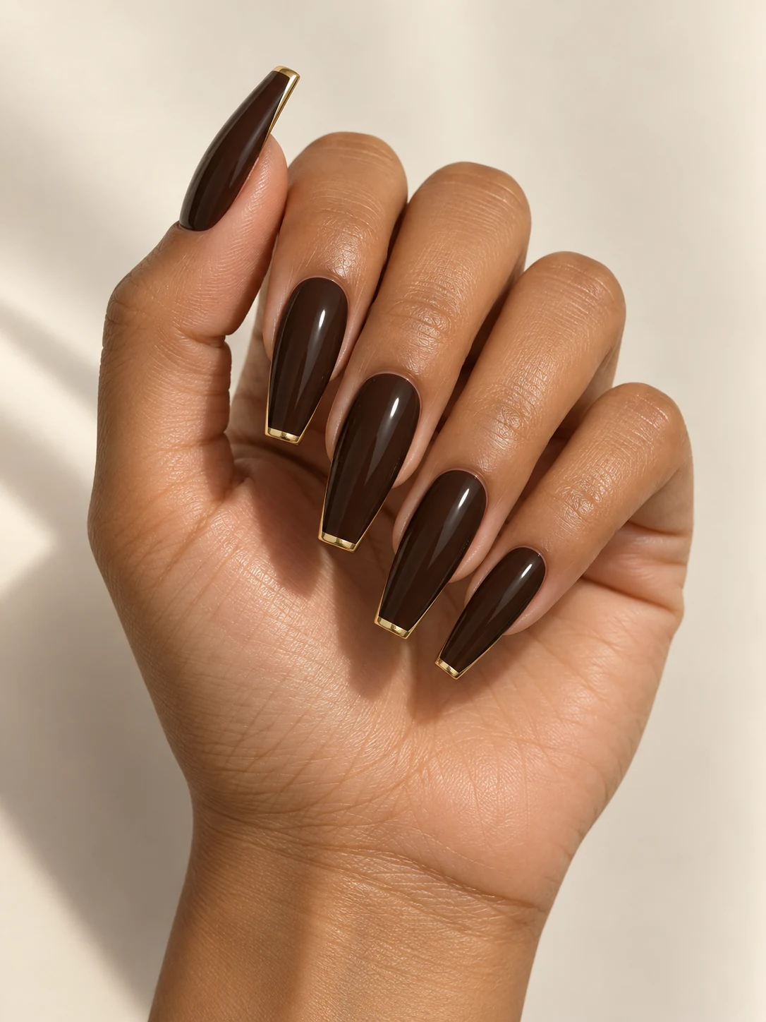 Deep espresso-brown coffin nails with razor-thin metallic gold French tips, rich lacquered finish on warm medium skin; long coffin manicure.
