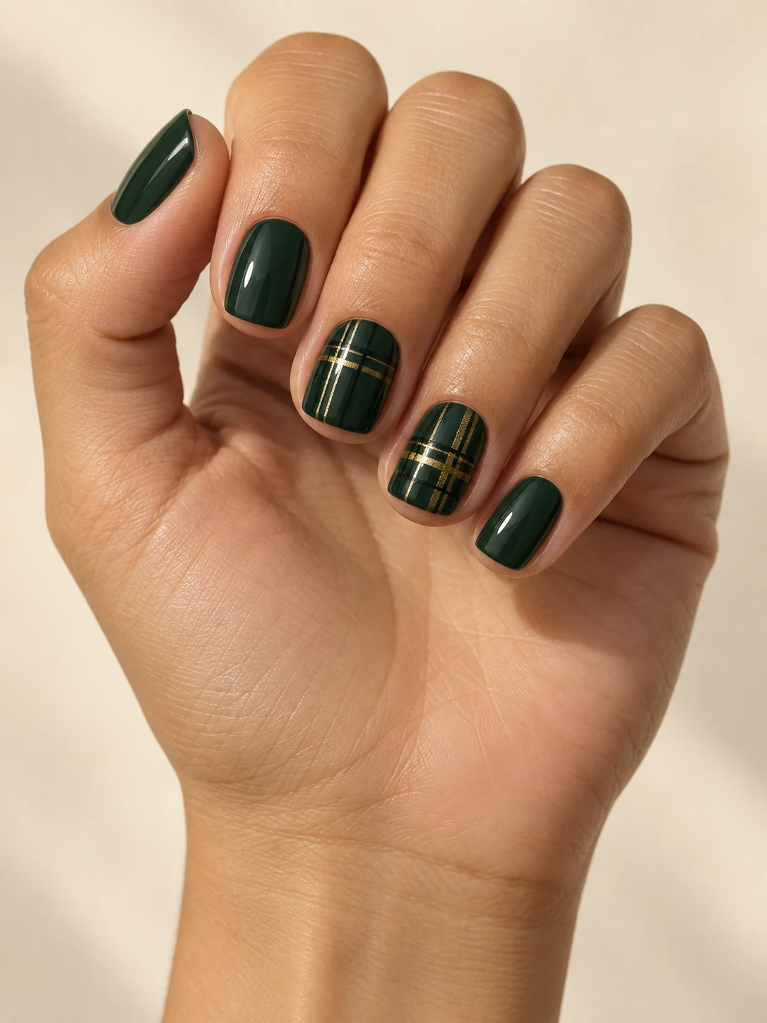 Deep forest green nails with fine black and gold tartan lines crossing two accent nails only on light golden skin; short square manicure.