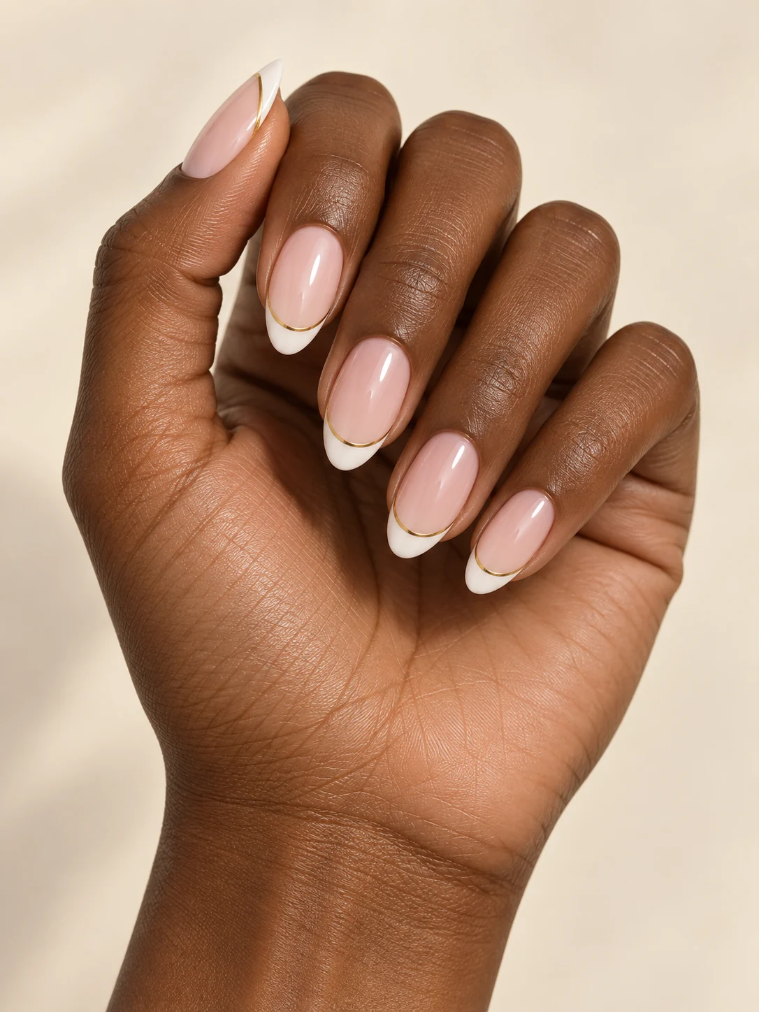 Milky pink nails with thin white French tips bordered below by a single delicate metallic gold thread on deep warm brown skin; medium almond manicure.