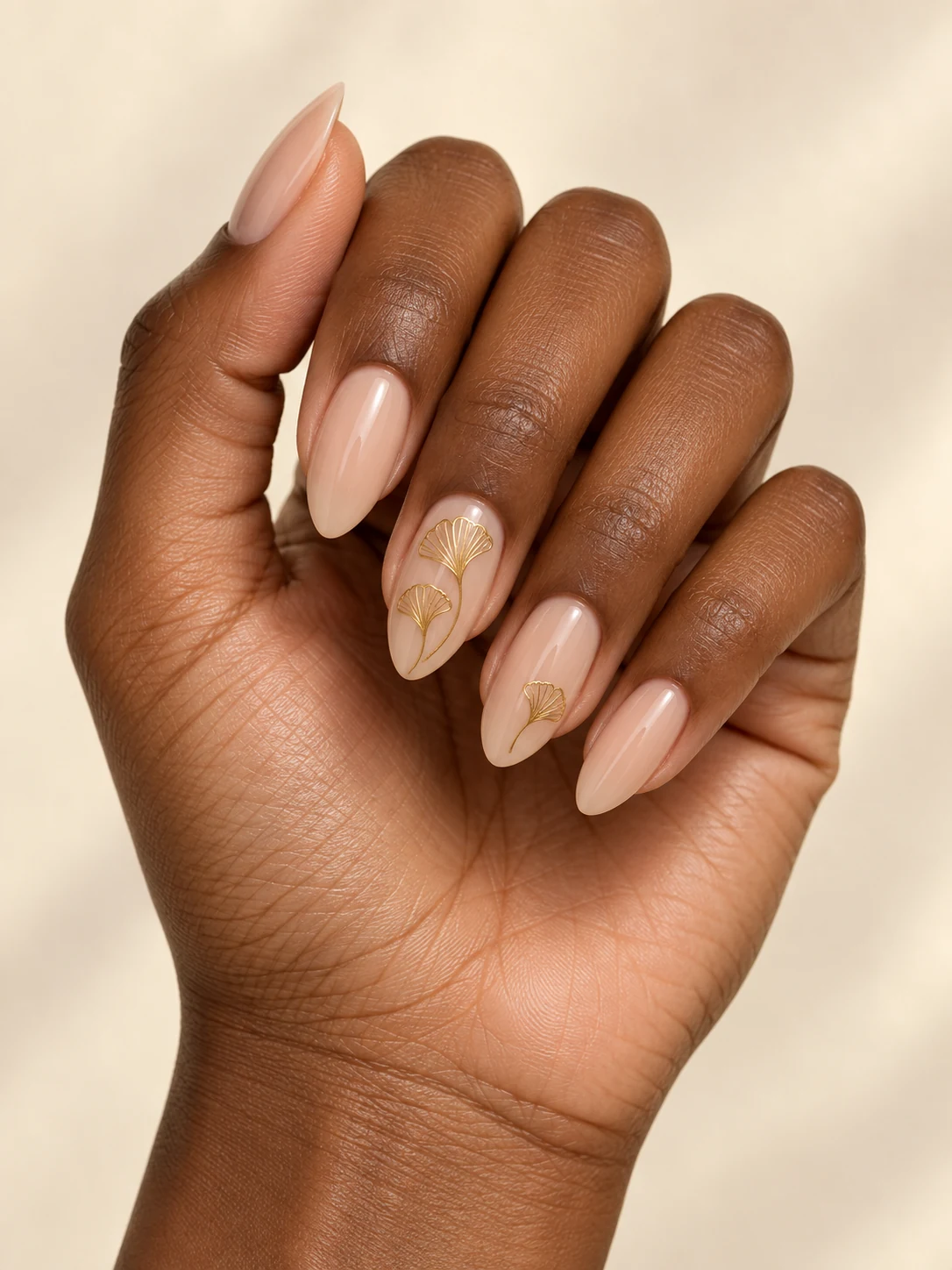 Sheer warm beige nails with fine fan-shaped golden ginkgo leaf outlines on two accent nails, no solid gold fill on rich deep brown skin; medium almond manicure.