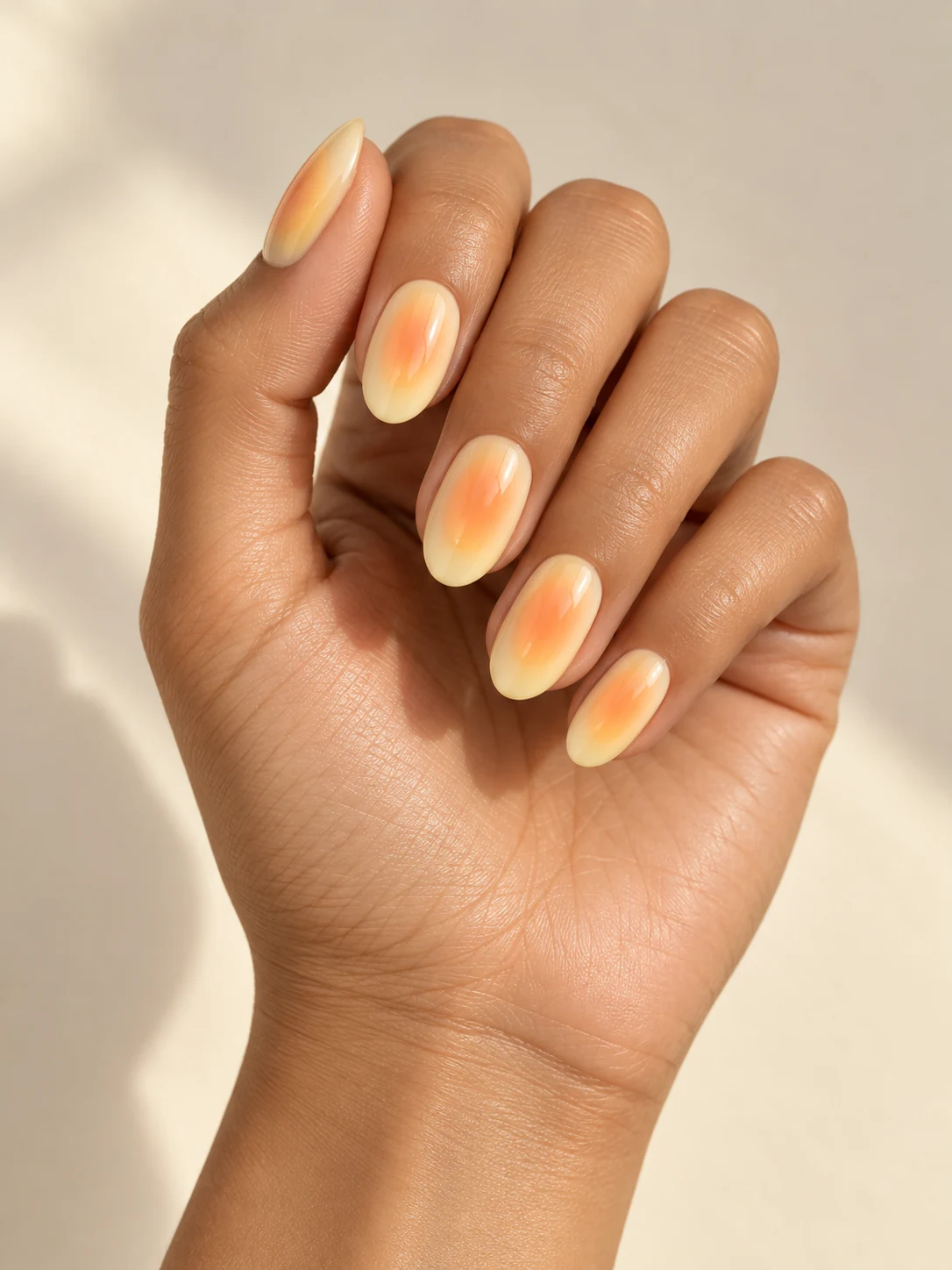 Soft pale yellow oval nails with glowing peach-orange aura centers, diffused sunlit color and glossy finish on warm medium skin; medium oval manicure.