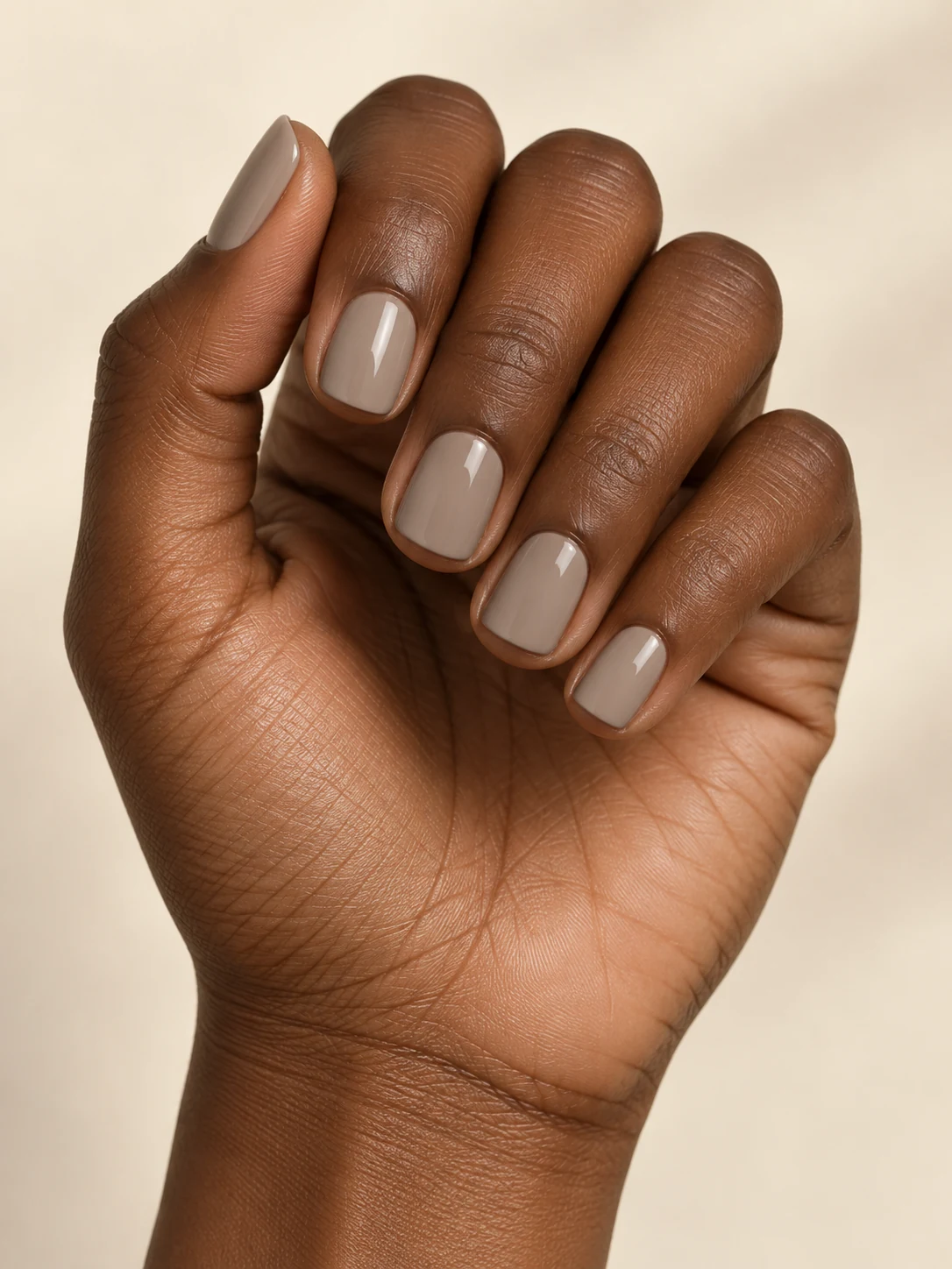 Short square nails in a cool mushroom greige cream, understated soft glossy finish and no pattern on deep warm brown skin; short square manicure.