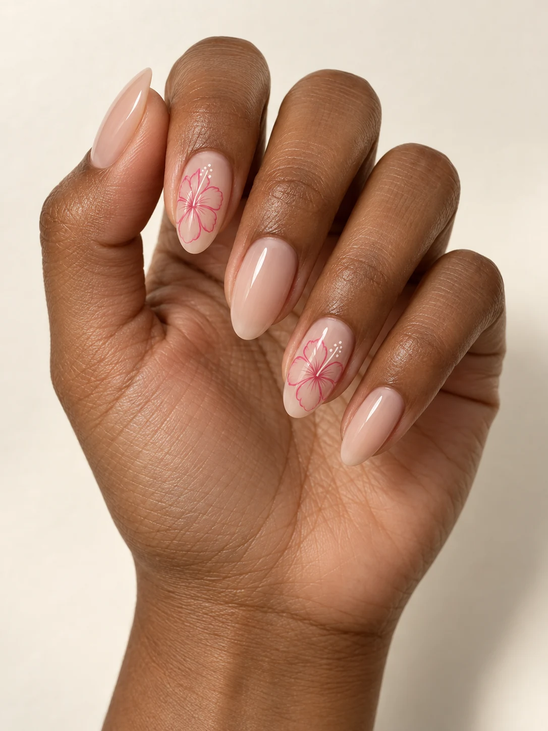 Nude almond nails with a fine pink single-line hibiscus bloom on two accent nails, white dot center and airy unpainted space on deep warm brown skin; medium almond manicure.