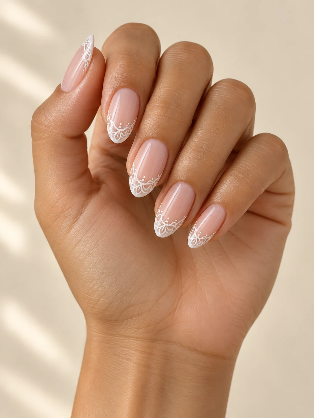 Sheer blush nails with delicate flat white lace-like loops restricted to the free edges, airy scalloped bridal detail on olive tan skin; medium almond manicure.