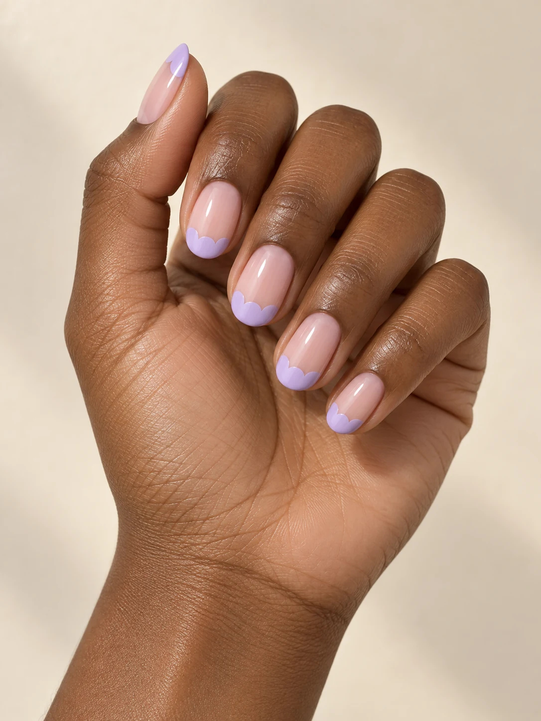 A nude pink base with soft lavender scalloped French tips, three rounded scallops along each free edge on deep warm brown skin; medium oval manicure.