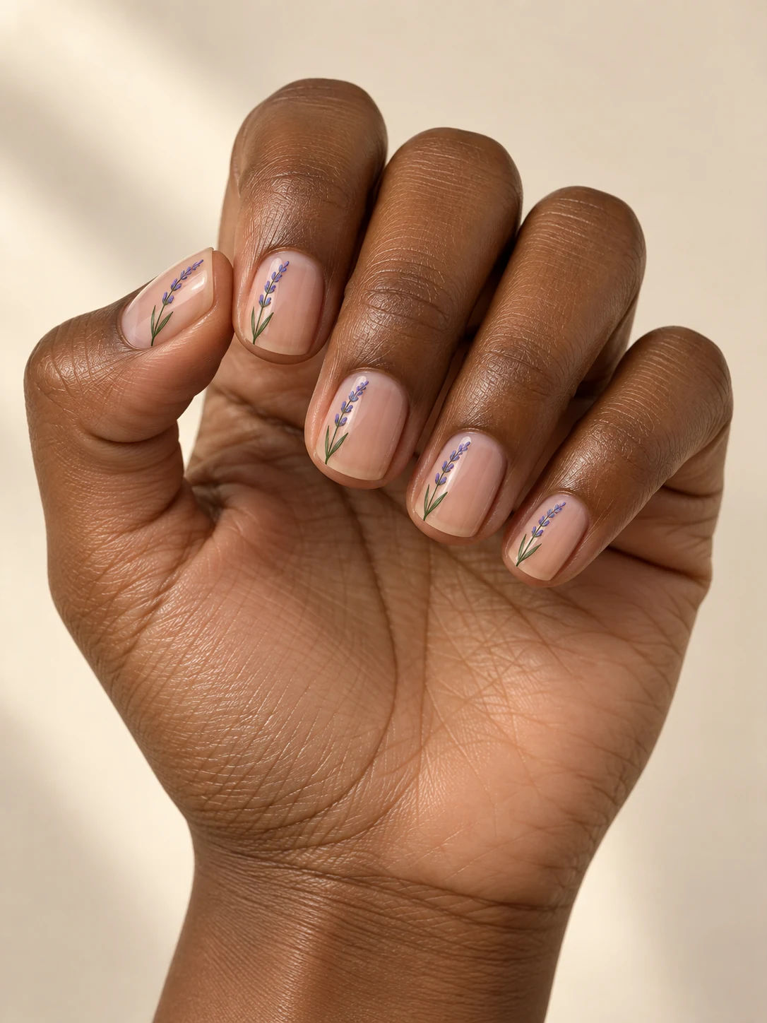 Clear nude short square nails with thin green lavender stems and tiny violet buds placed vertically near one side on deep warm brown skin; short square manicure.