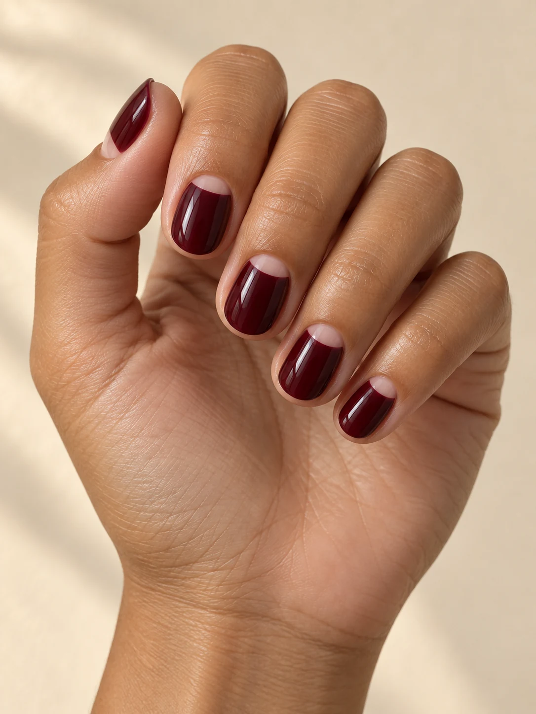 Glossy deep merlot nails with clean small bare-looking semicircular half moons at the cuticle on olive tan skin; short squoval manicure.