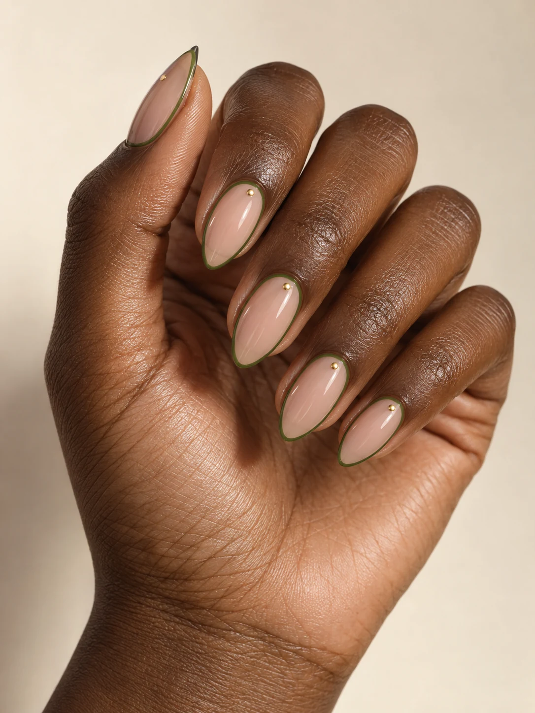 A sheer warm nude center enclosed by a fine olive-green perimeter, with one tiny gold dot near each cuticle on warm medium skin; medium almond manicure.
