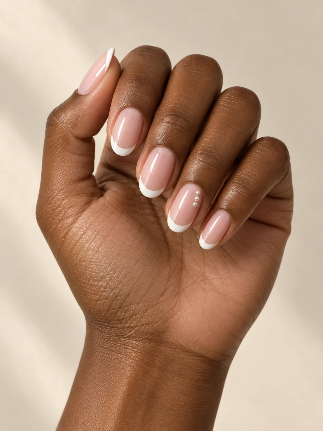 Milky pink nails with white French tips and a tiny row of three white pearl-like dots on one ring-finger accent on deep warm brown skin; medium oval manicure.