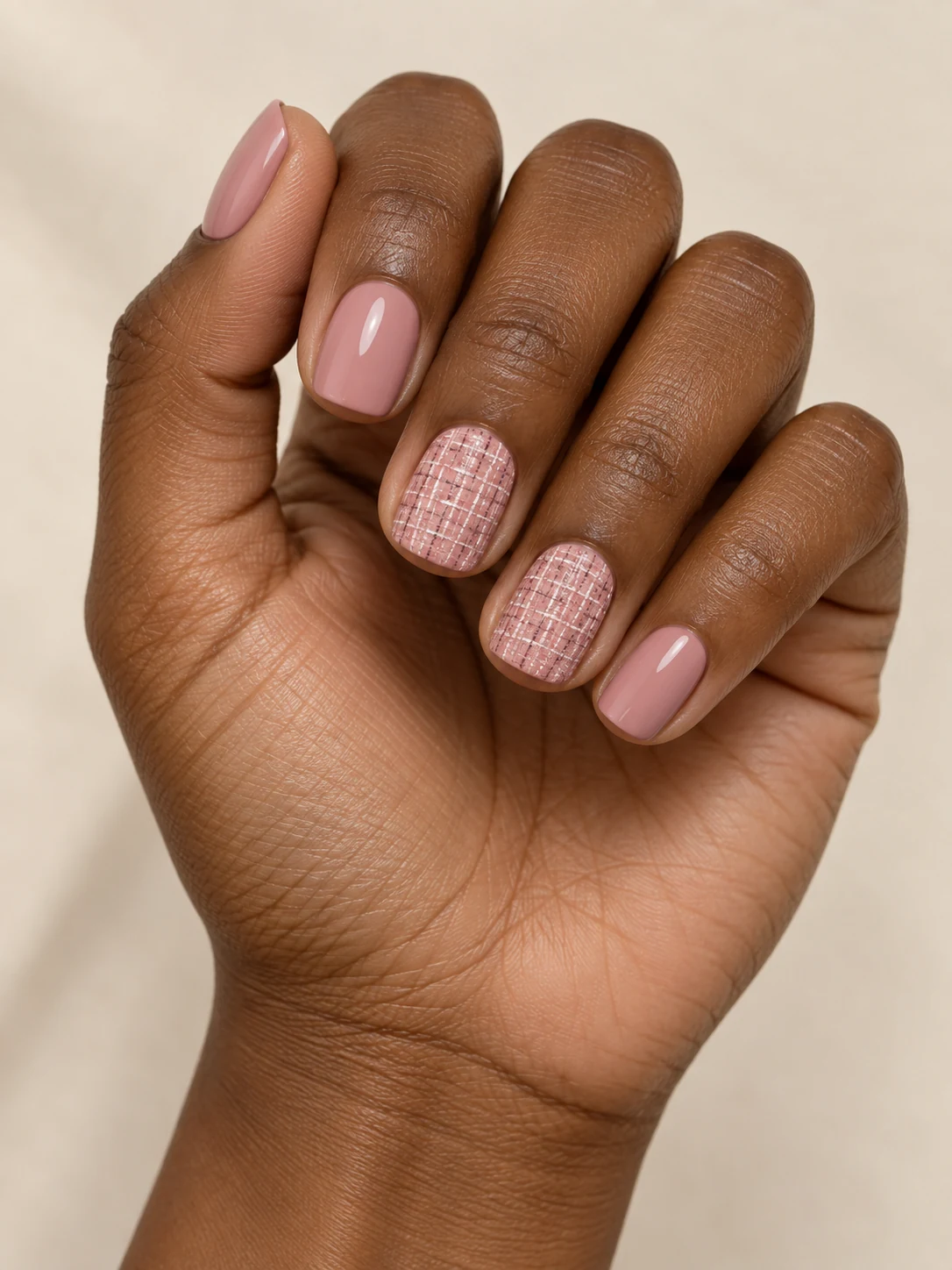 Dusty pink nails with fine intersecting ivory and deeper pink tweed-like woven strokes on two accent nails only on deep warm brown skin; short squoval manicure.