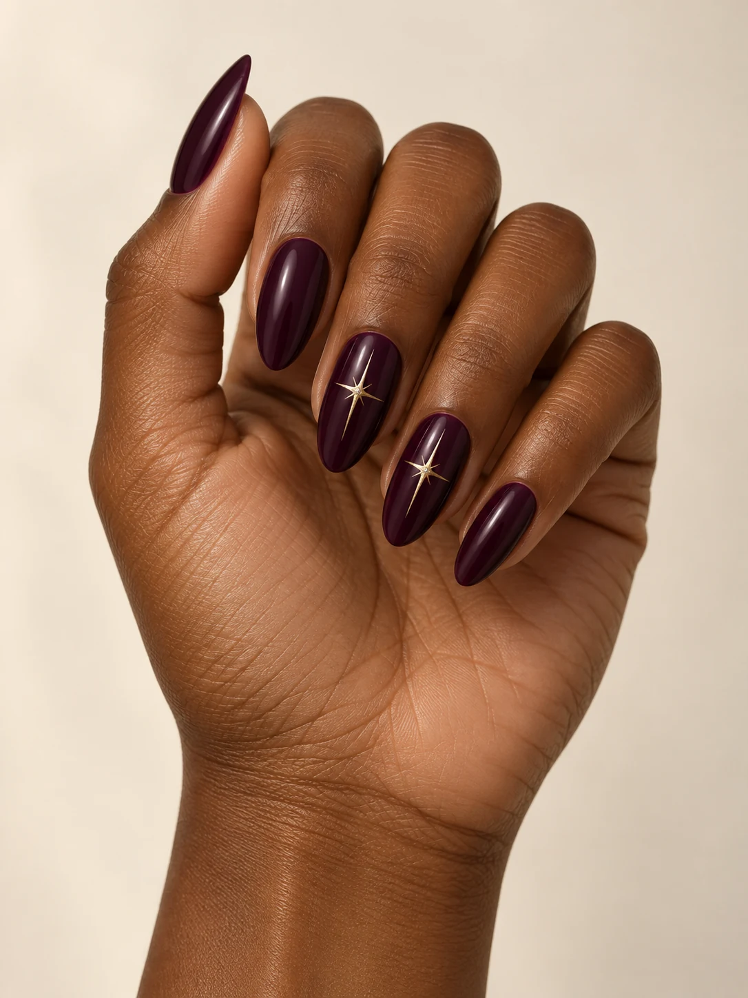 Deep plum nails with one delicate gold starburst on each ring and middle nail, remaining nails plain plum on deep warm brown skin; long almond manicure.
