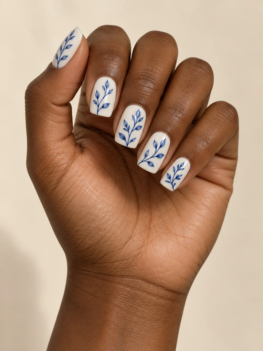 Ivory-white nails with fine cobalt-blue botanical stems and small leaves, delicate hand-painted porcelain style on deep warm brown skin; medium square manicure.