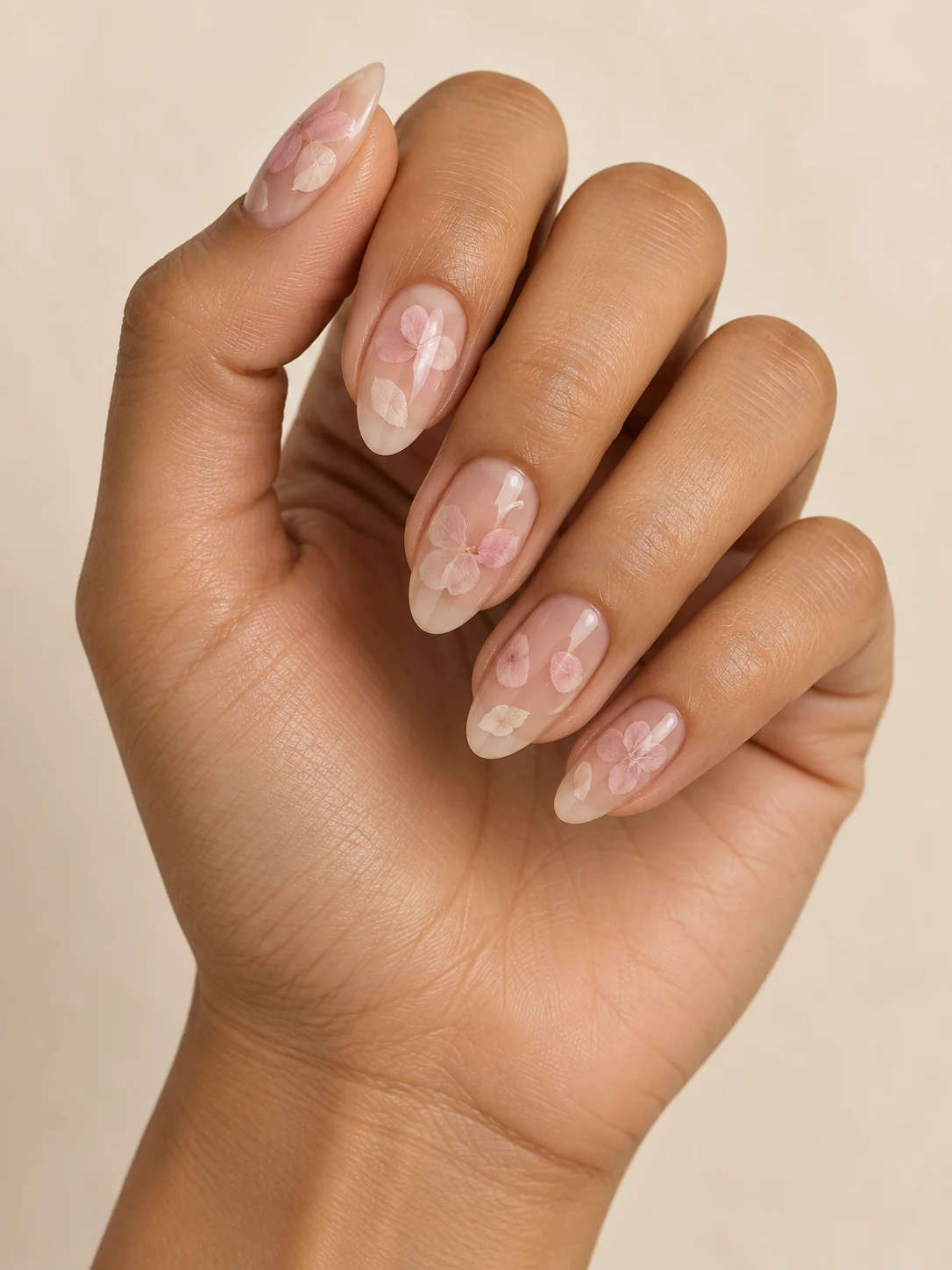 Translucent nude nails with delicate small pale pink and ivory petal silhouettes appearing suspended beneath clear gloss on warm medium skin; medium almond manicure.