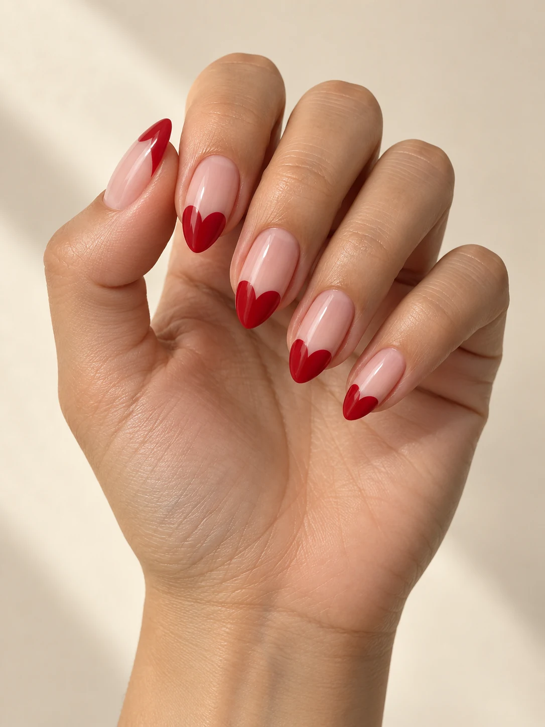 Nude almond nails with red heart-shaped French tips, two rounded red lobes meeting toward each tapered tip on light golden skin; medium almond manicure.