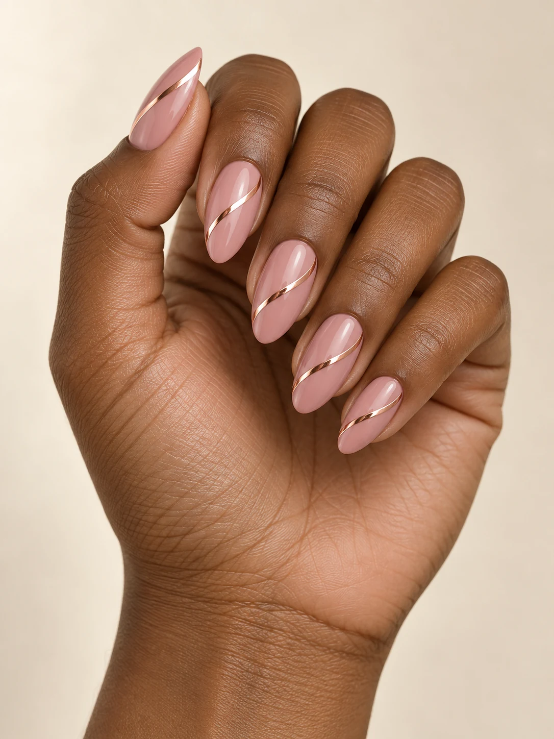 Dusty pink almond nails with one thin metallic rose-gold ribbon spiraling diagonally across each nail, no full chrome coverage on deep warm brown skin; medium almond manicure.