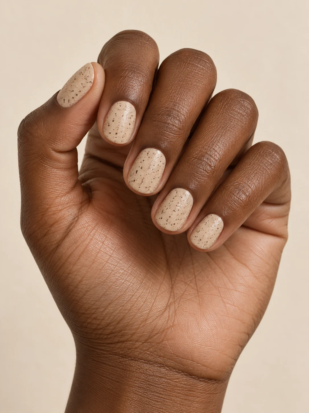 Creamy sand-beige nails sprinkled with extremely fine brown stone-like speckles, matte-satin surface on rich deep brown skin; short round manicure.