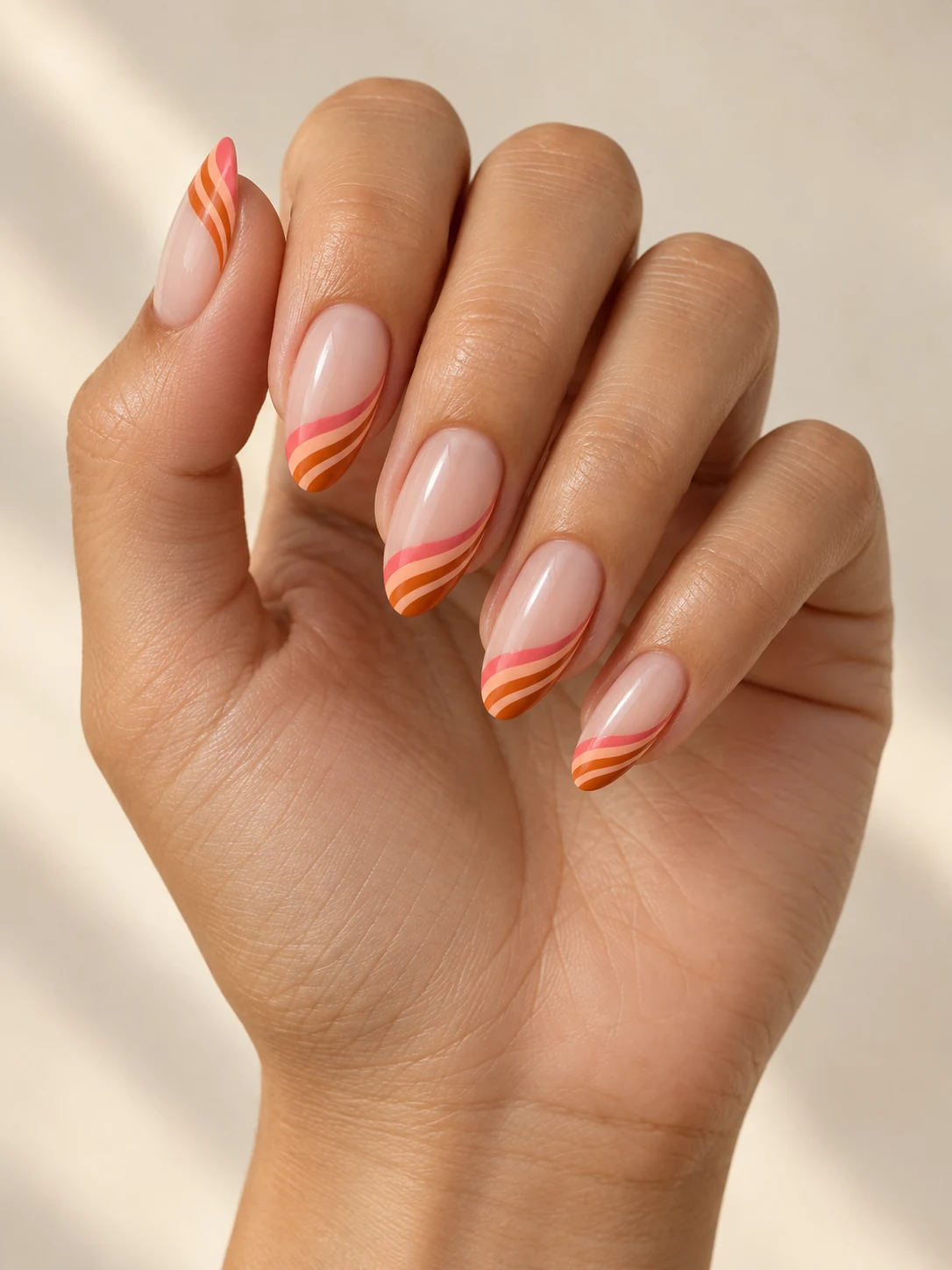 Sheer nude nails with three slim curved stripes in coral pink, peach and burnt orange near each free edge on light golden skin; medium almond manicure.