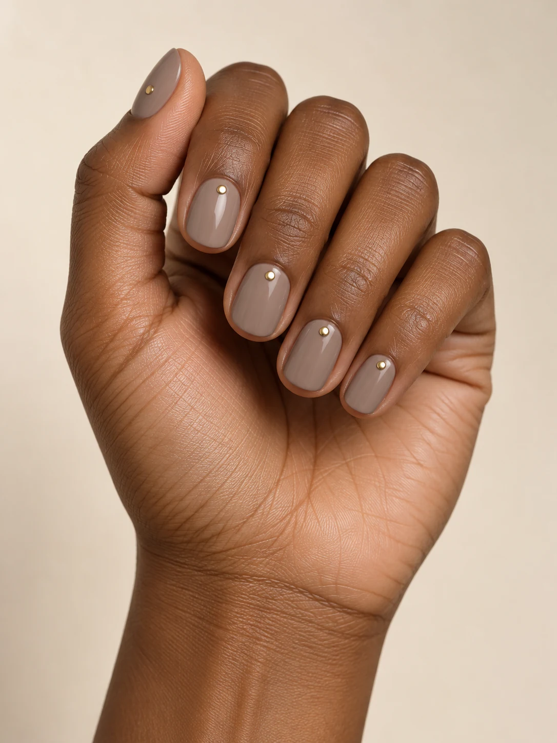 Taupe cream nails with one minute gold dot centered just above each cuticle, muted glossy office style on deep warm brown skin; short round manicure.