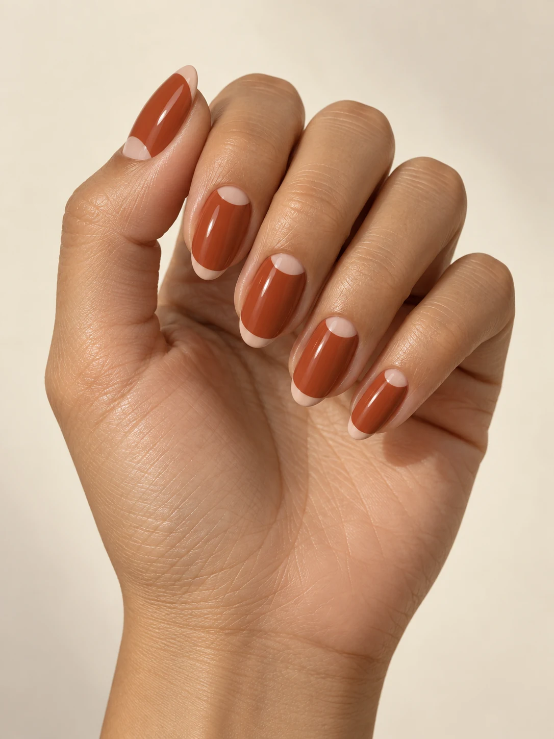 Warm terracotta nails with small nude half moons near the cuticle and a narrow nude semicircle at the free edge on light golden skin; medium oval manicure.