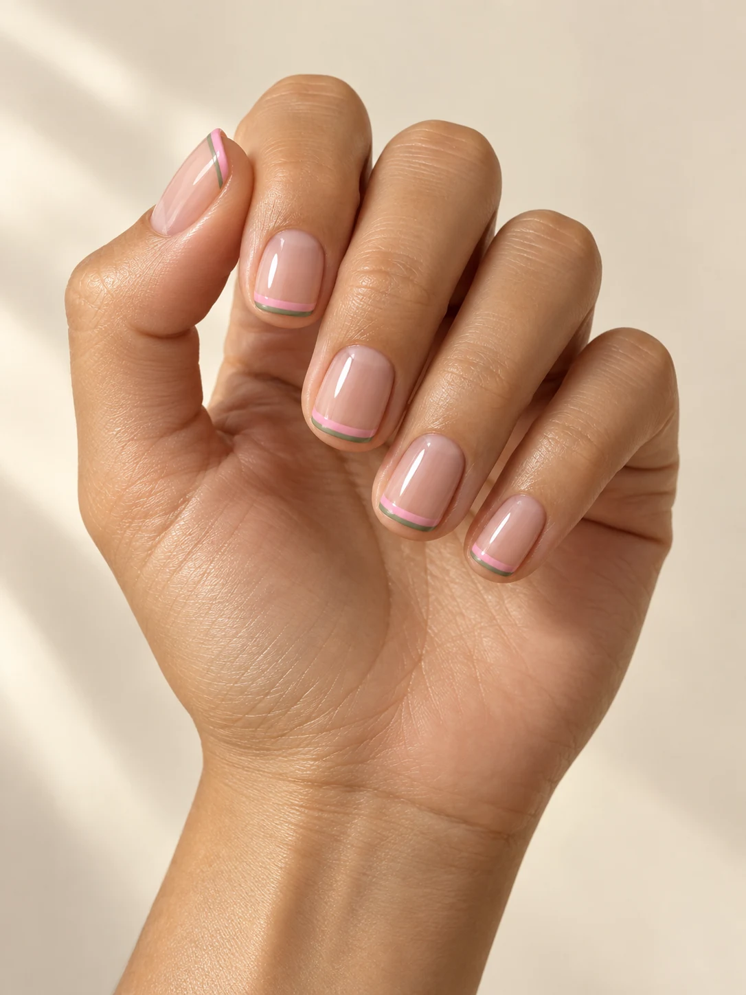 Sheer nude short square nails with a narrow pink stripe and an even thinner muted green stripe tracing each free edge on light golden skin; short square manicure.