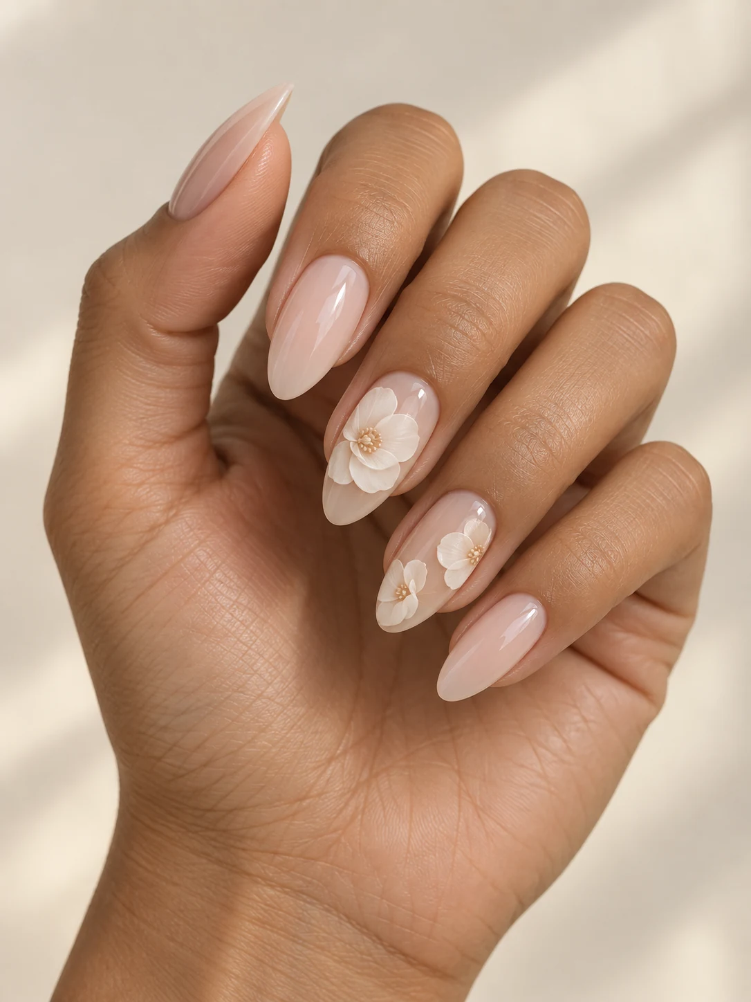 Milky nude nails with layered-looking ivory camellia petals clustered on two accent nails, with the remaining nails understated nude on olive tan skin; long almond manicure.
