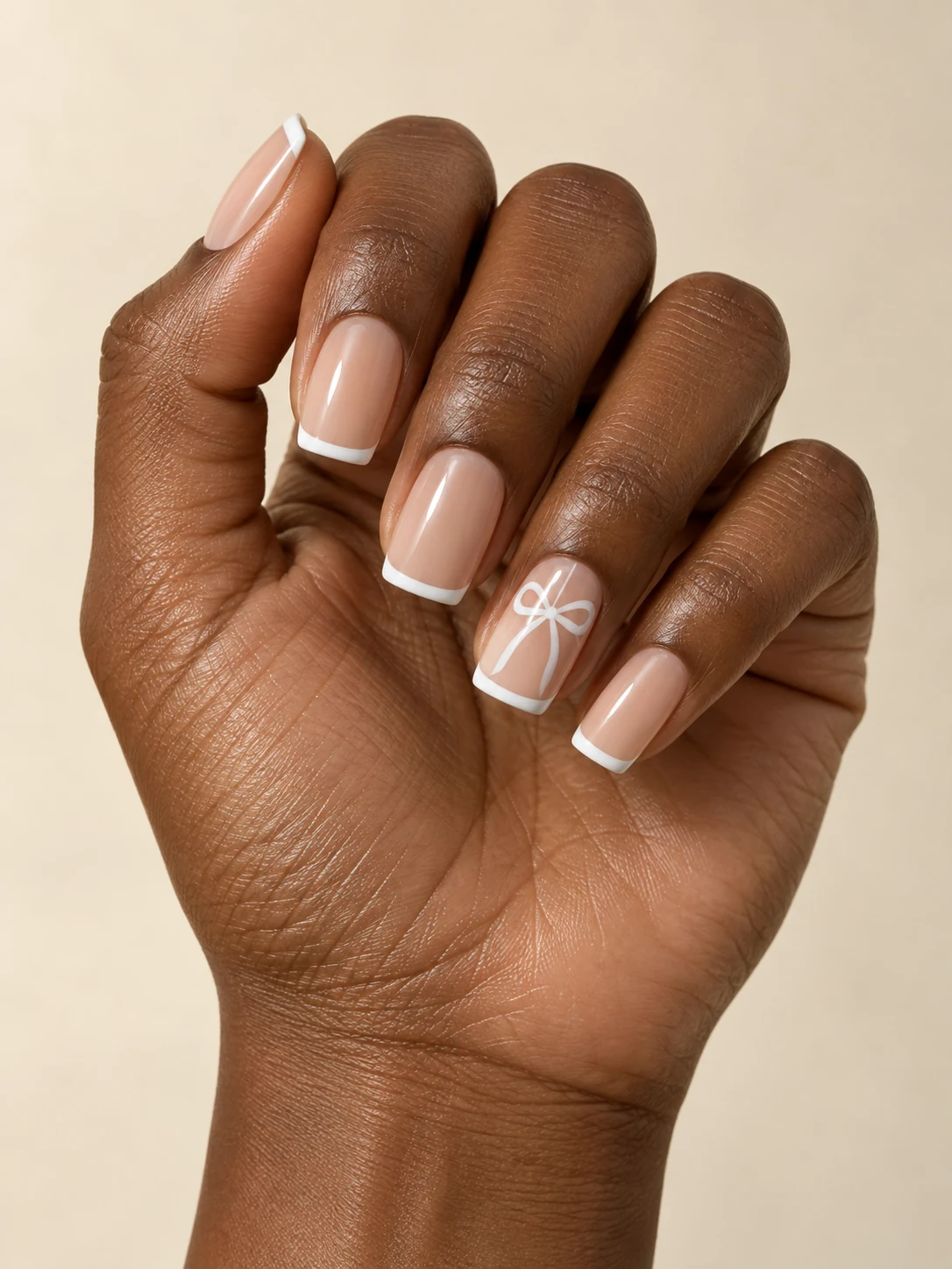 Sheer beige square nails with a slim flat white ribbon bow drawn on each ring-finger nail and clean white micro tips on rich deep brown skin; medium square manicure.